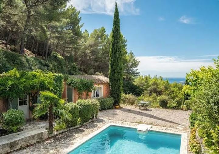 Piscine chauffée de la Villa L’Amalié entourée de verdure, avec la maison, un grand cyprès et la mer à l’horizon